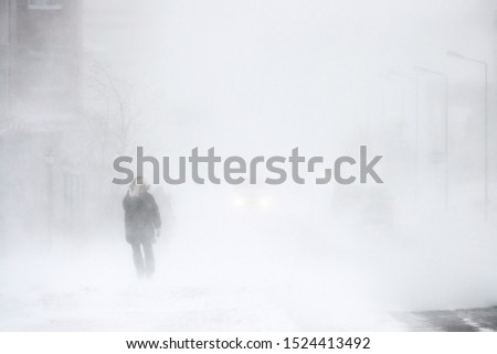 Snowstorm in the city. A man during a blizzard is walking along the street. Cars on a snowy road. Strong wind and snowfall. Arctic climate. Extreme North. Anadyr, Chukotka, Siberia, Far East Russia. Royalty-Free Stock Photo #1524413492