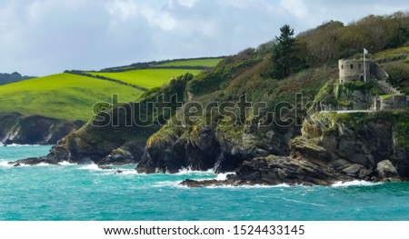 Landscape of the River Fowey estuary, from Polruan. Distant views of rugged cliffs, tree lined hills, fields and a 16th. Century fortification. Blue sea with white surf breakers. Cornwall. England.