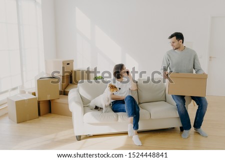 Photo of happy couple move in new home, pose on sofa with pet and boxes, have relocation, unpack boxes, female calls delivery service, carry belongings in carton containers. Welcome to new apartment Royalty-Free Stock Photo #1524448841