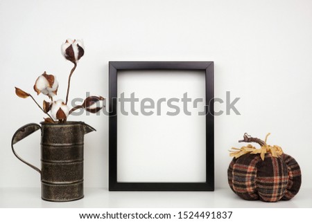 Mock up black frame with cotton branch and plaid pumpkin decor on a shelf or desk. Autumn concept. Portrait frame against a white wall.