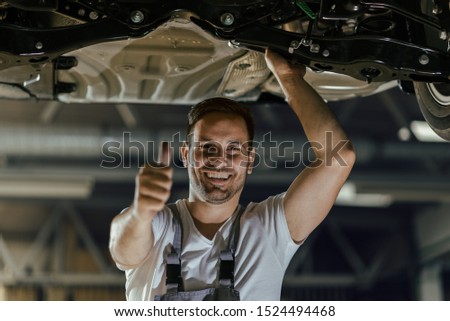 Happy mechanic showing thumbs up while examining undercarriage of a car in a workshop. He is looking at camera Royalty-Free Stock Photo #1524494468