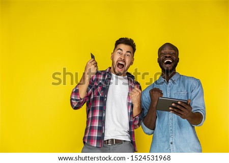 european and afroamerican guy are sincerely exited, looking in front of with the tablet and credit card in hands on the yellow background