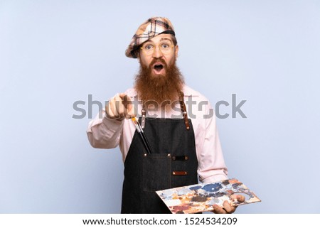 Redhead artist man with long beard holding a palette over isolated blue background surprised and pointing front