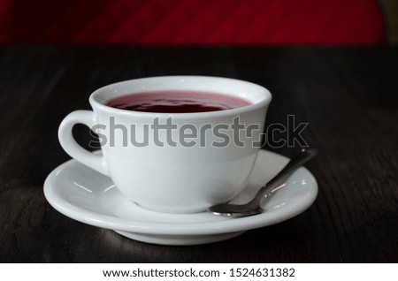 A white cup on a white saucer and spoon, with red tea, on dark wooden table in a dark cafe closeup