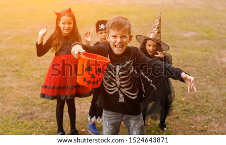 Spooky little boy in skeleton costume holding trick or treat bucket and intimidating camera while having fun with friends during Halloween party in park