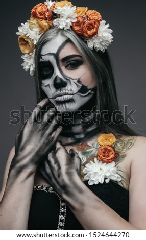 Young woman with spooky makeup and in flower wreath touching hair and looking at camera during Dia de Muertos party against gray background