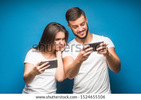 Image of unhappy woman and man frowning and peeking at each others cell phones, isolated over blue background.