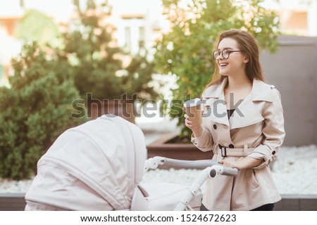 Stylish young woman holding disposable coffee cup and looking at baby stroller.