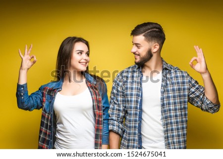Portrait of content couple in basic clothing smiling at camera while doing okay sign, isolated over yellow background. Lovely concept.