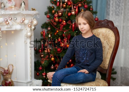 Barefoot young girl wearing a blue long sleeve shirt and jeans sitting in a chair. Fireplace filled with toys and decorated Christmas tree background.