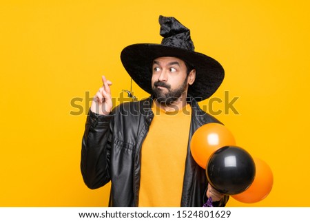 Man with witch hat holding black and orange air balloons for halloween party with fingers crossing and wishing the best