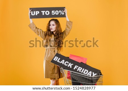 young woman in trench coat with black friday 50% sign and colorful shopping bags in cart isolated over yellow