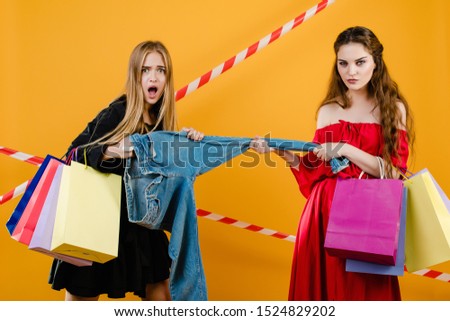 young women wearing dresses fighting over pair of jeans with shopping bags isolated over yellow