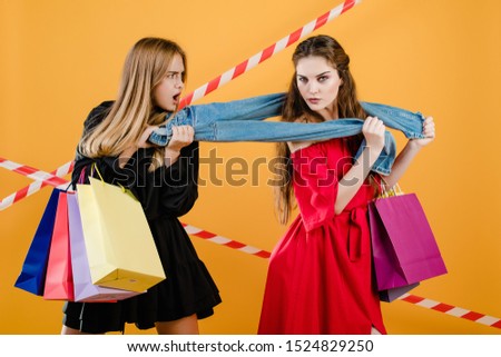 young women wearing dresses fighting over pair of jeans with shopping bags isolated over yellow