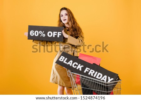 smiling woman in autumn coat with black friday 60% sign and colorful shopping bags in cart isolated over yellow