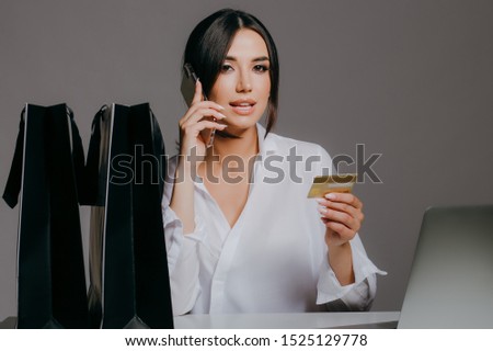 A beautiful girl in a white shirt, with a laptop, looking at credit card and holding a phone, preparing for black Friday isolated on grey