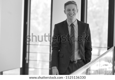 Black and white photo of smiling young businessman standing against window at office