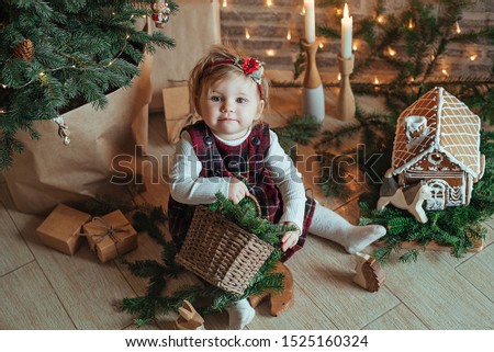 Cute little girl is sitting by the Christmas tree on the floor in the room. Next to the child is a gingerbread gingerbread house and a basket with fir branches. Merry christmas concept. Cozy  home