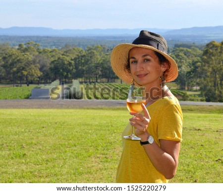 A young beautiful smiling woman wearing a hat and holding a glass cold white wine admiring a beautiful landscape of winery fields, Hunter Valley, Australia