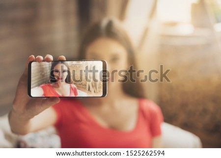 Young woman taking selfie using phone. Sit on sofa in room alone. Prepare for good shot. Smartphone screen. Daylight in room. Modern technologies