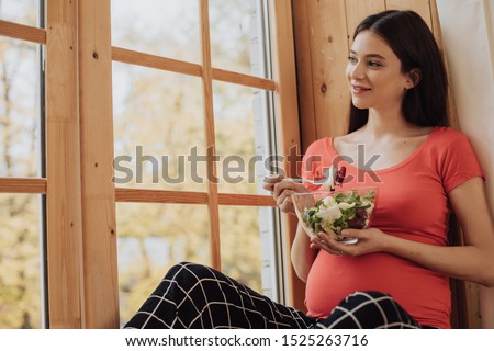 Bautiful picture of nice young pregnant woman sitting at window and eat salad from glass bowl. Look at windown and smile. Relaxed calm and peaceful brunette. Maternity time. Future single mom
