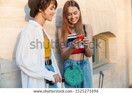 Photo of two happy tourist women holding cellphone while walking with suitcases on city street