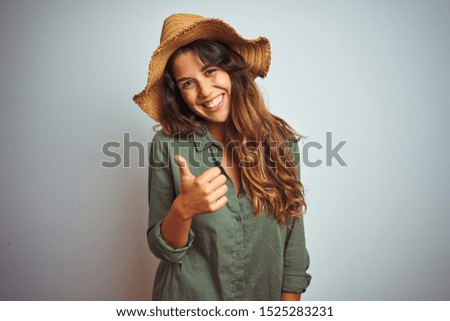 Young beautiful woman on vacation wearing green shirt and hat over white isolated background doing happy thumbs up gesture with hand. Approving expression looking at the camera with showing success.