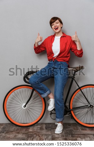 Full length image of beautiful woman showing thumb up and standing by stylish bicycle isolated over gray background