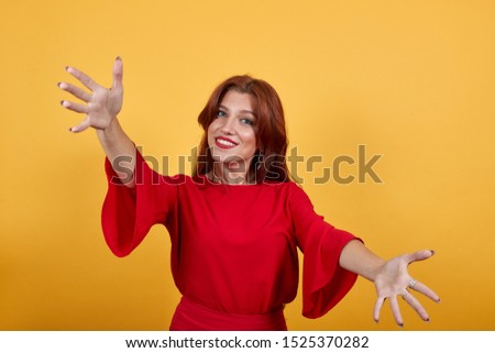 Happy girl smiling, holding hands widely opened and shows gesture of open arms. Woman in delightful dress stands on orange background. 