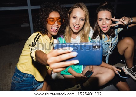 Image of a young cheery girls friends at night on street take a selfie mobile phone.