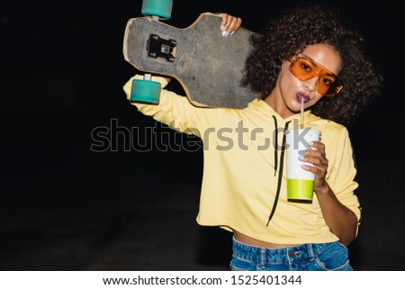 Image of glamour african american girl in streetwear drinking soda and holding skateboard at night outdoors