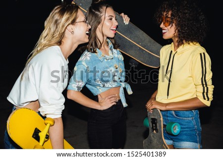 Image of positive multiethnic girls in streetwear smiling and holding skateboards at night outdoors