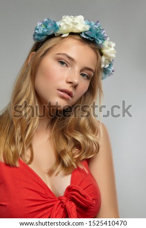 Close-up shot of a romantic blonde European lady with wavy hair in a fire-red low necked top and a delicate circlet made of white and dusky blue artificial flowers.  