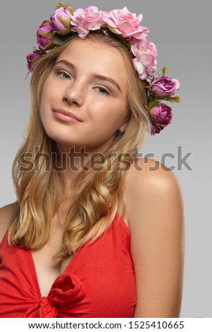 Close-up shot of a romantic blonde European lady with wavy hair in a fire-red low necked top and a delicate circlet made of pink and purple artificial roses.  