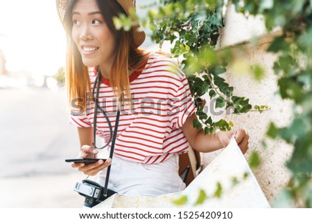 Photo of a young smiling positive beautiful asian woman walking outdoors with camera holding map using mobile phone.
