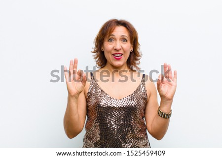 middle age pretty woman feeling shocked, amazed and surprised, showing approval making okay sign with both hands against white wall