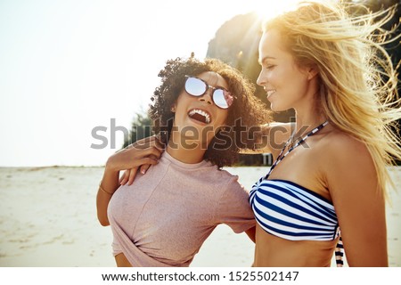 Two laughing young women walking arm in arm together along a tropical beach while on summer vacation