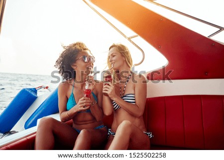 Two smiling young female friends wearing bikinis sitting together on a boat on the open ocean during summer vacation sipping on drinks