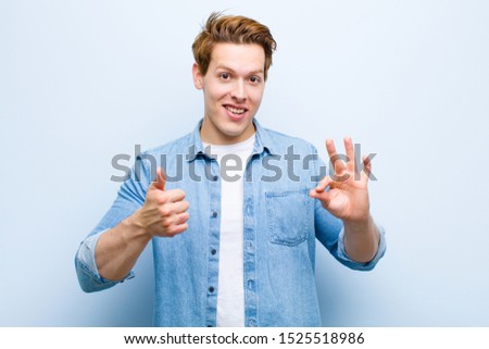 young red head man feeling happy, amazed, satisfied and surprised, showing okay and thumbs up gestures, smiling against blue wall