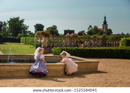 Cute blond curly twin girls dressed up as princesses in the garden of ancient castle, beautiful small girls in blue and pink long dresses, fairy tale scene in spring