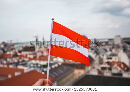 A man holds in his hand the flag of Austria on a viewing platform of St. Stephen's Cathedral in Vienna against the backdrop of a beautiful sunset sky and city panorama.