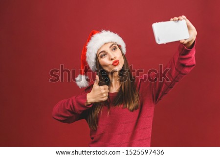 Christmas portrait of beautiful girl. Teenager wearing santa claus hat. Girl smiling and making selfie on mobile phone isolated against red background. 