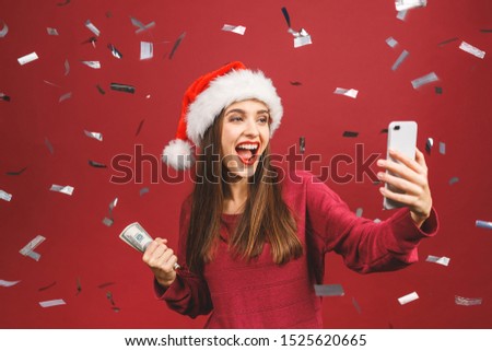 Happy winner! Pensive brunette woman in christmas hat holding money isolated over red background. Selfie concept. Using phone. 