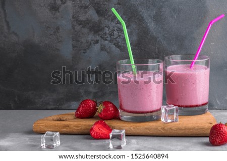 two glasses of strawberry cocktail with straws on a wooden board and gray background with ice cubes, refreshment drinks for summer, copy space fot text
