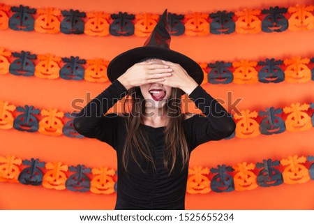 Image of mystic witch girl in black halloween costume covering her eyes and smiling isolated over orange pumpkin wall