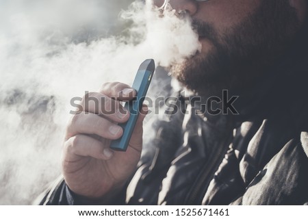 Man smokes new Vape Pod System, inhales and exhales vapor of electronic cigarette, vaping concept, selective focus, toned Royalty-Free Stock Photo #1525671461