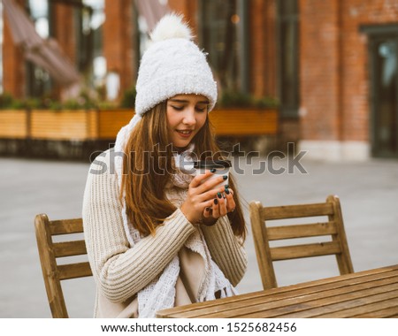 Beautiful young girl drinking coffee, tea from plastic mug in autumn, winter. A woman with long hair in warm clothes sitting in street cafe, warmed by a hot drink