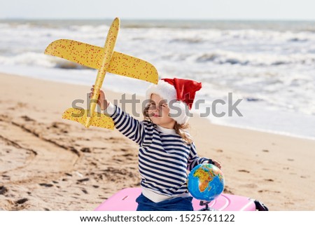 Little girl in Santa hat playing and having fun with yellow toy airplane on sunny tropical beach. Christmas and new year vacation travel concept