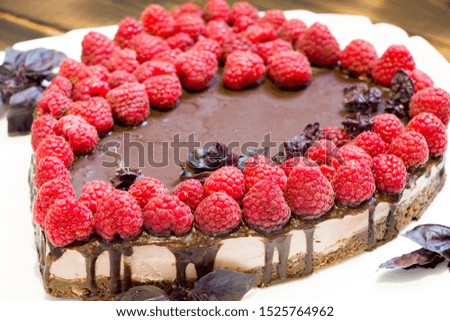 Close-up vegan cake topped with raspberries and chocolate on white plate with basil leaves. Cooked in the shape of a heart.