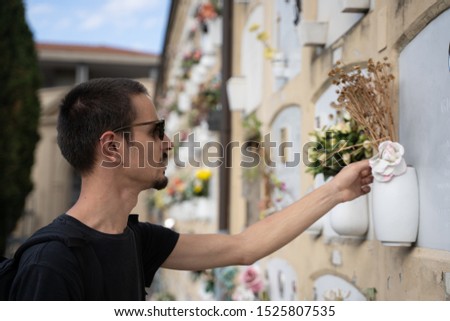 Stock photo of a boy dressed in black and with a suitcase and sunglasses, touching the flowers of a tomb in a cemetery in Bologna, Italy. Travel concept